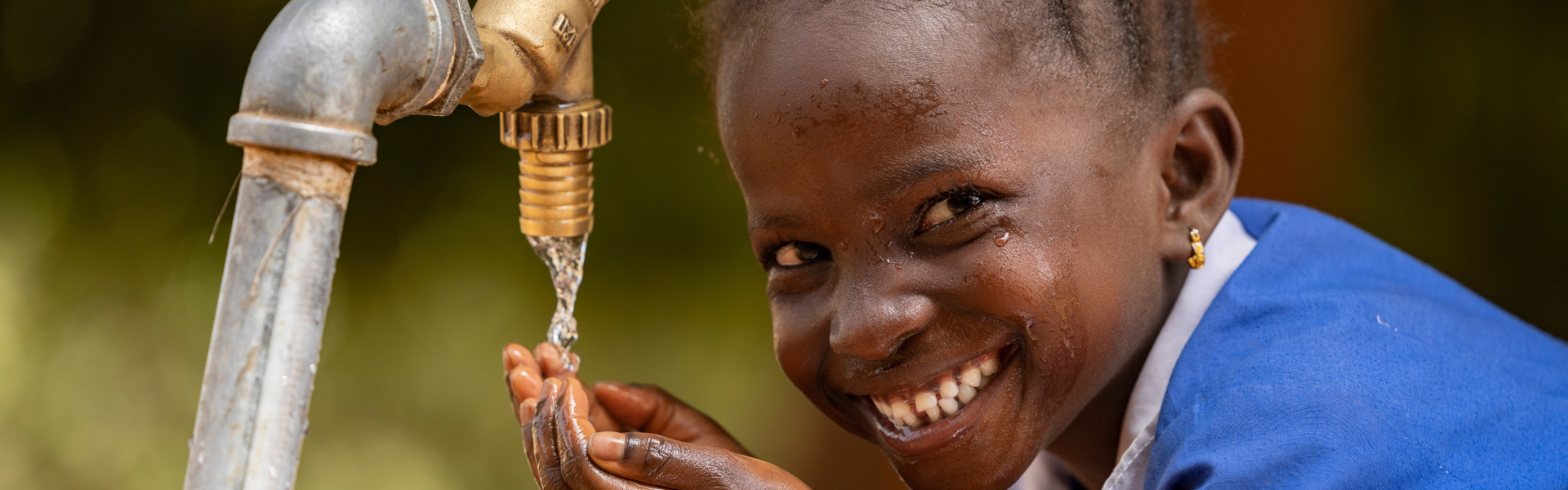 A little girl in a school uniform smiles at the camera while washing her face from an outdoor water faucet.