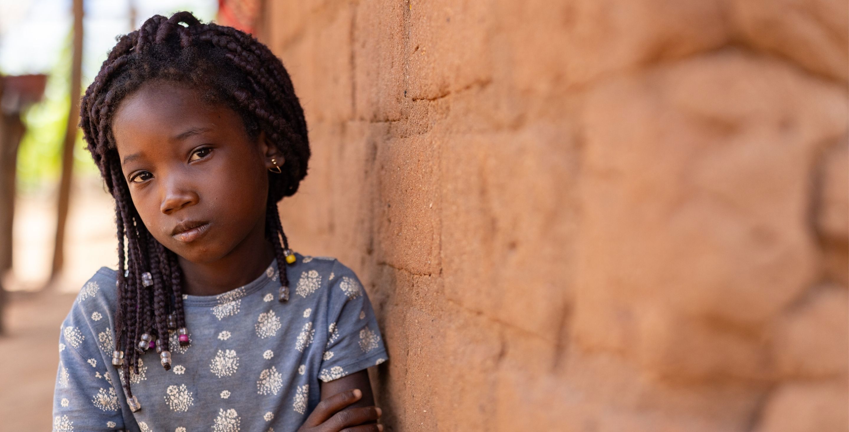A child in Mozambique sits against the wall of a hut, arms folded, looking pensively at the camera.
