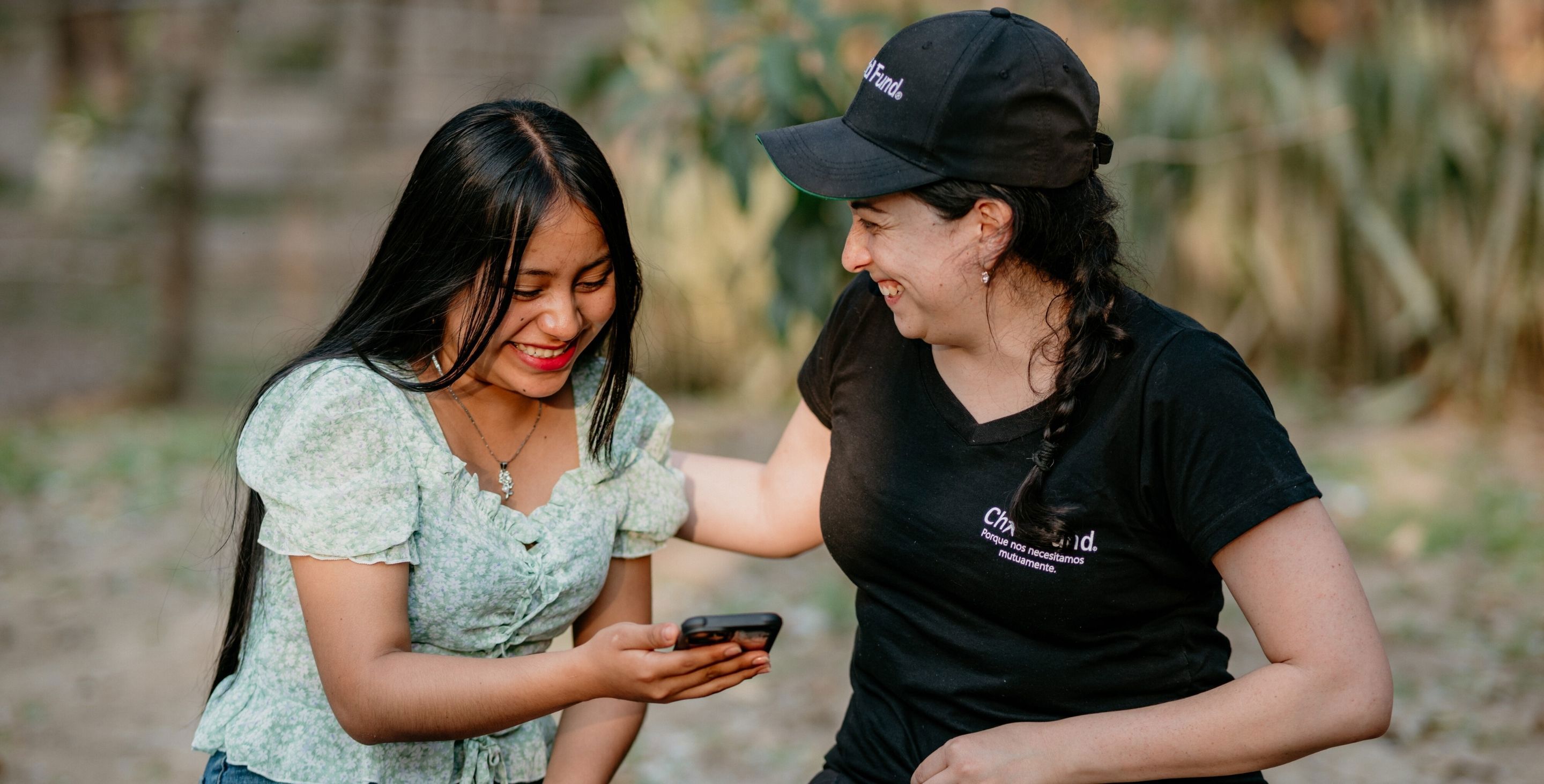 A teenage girl sits next to a woman in a ChildFund hat. Both are looking down at a cellphone and smiling.