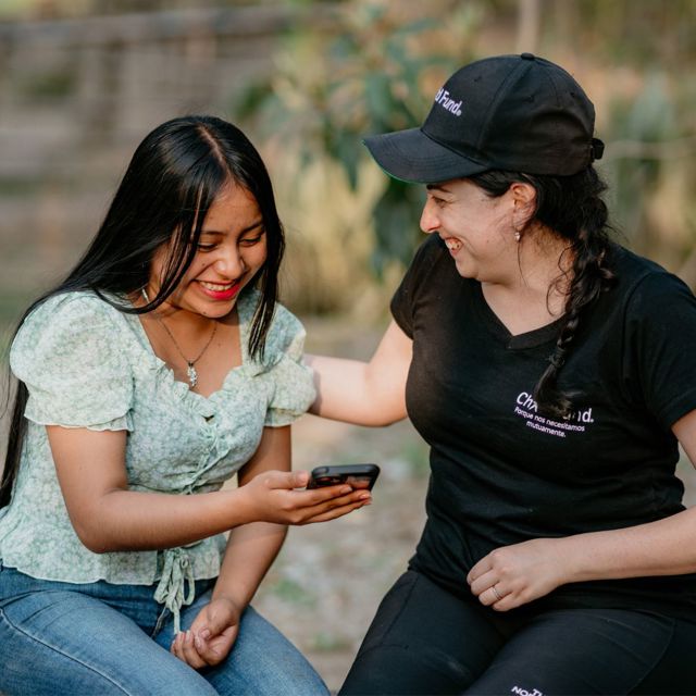 A woman in a ChildFund hat smiles at a teenage girl who is holding and looking down at her phone, smiling.