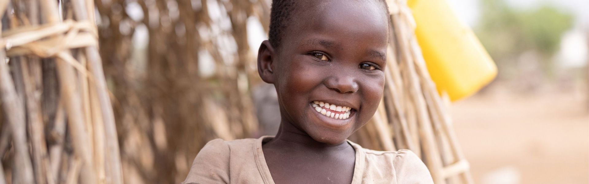 A girl in rural Kenya stands in front of a thatched hut, smiling at the camera.