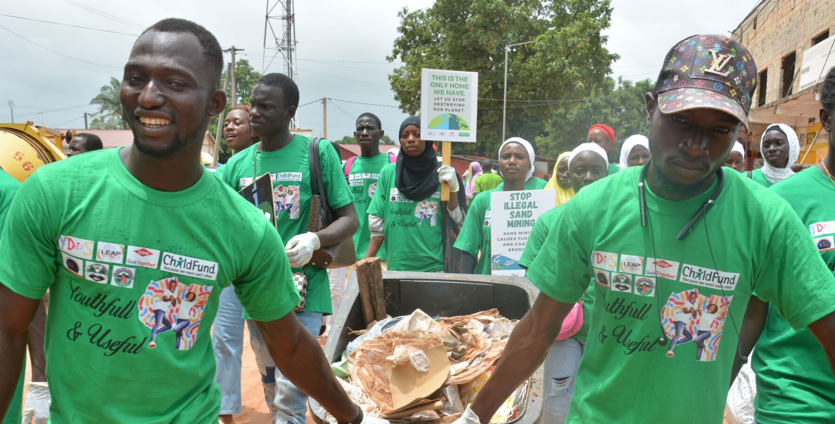 Young men parade down the street carrying a large garbage can, which they are filling with trash as they celebrate World Cleanup Day.