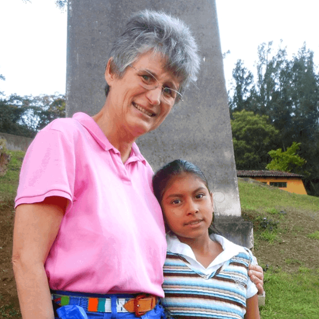 Susan Bush, a woman with short grey hair and glasses, wearing a pink polo shirt, stands with her arm around a young girl in a striped shirt. They are smiling together in front of a stone monument on a grassy hill.