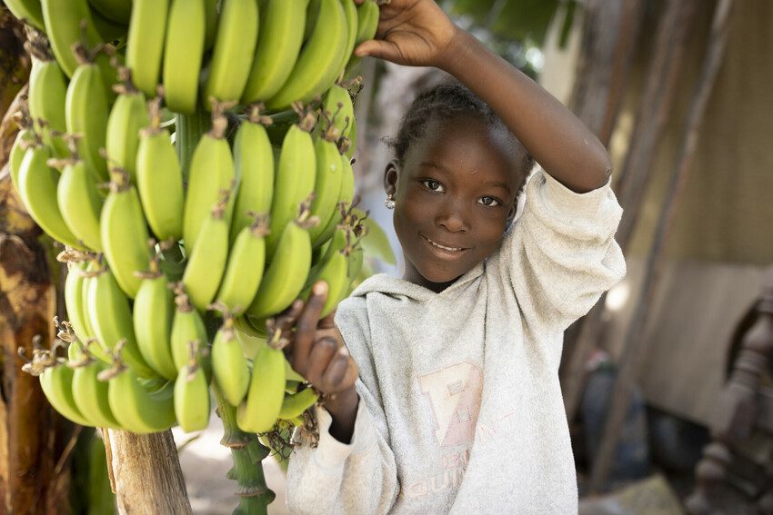 A young girl in Senegal smiles next to a bunch of bananas.