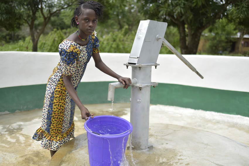 Girl stands outside at water pump well with blue bucket in Sierra Leone.