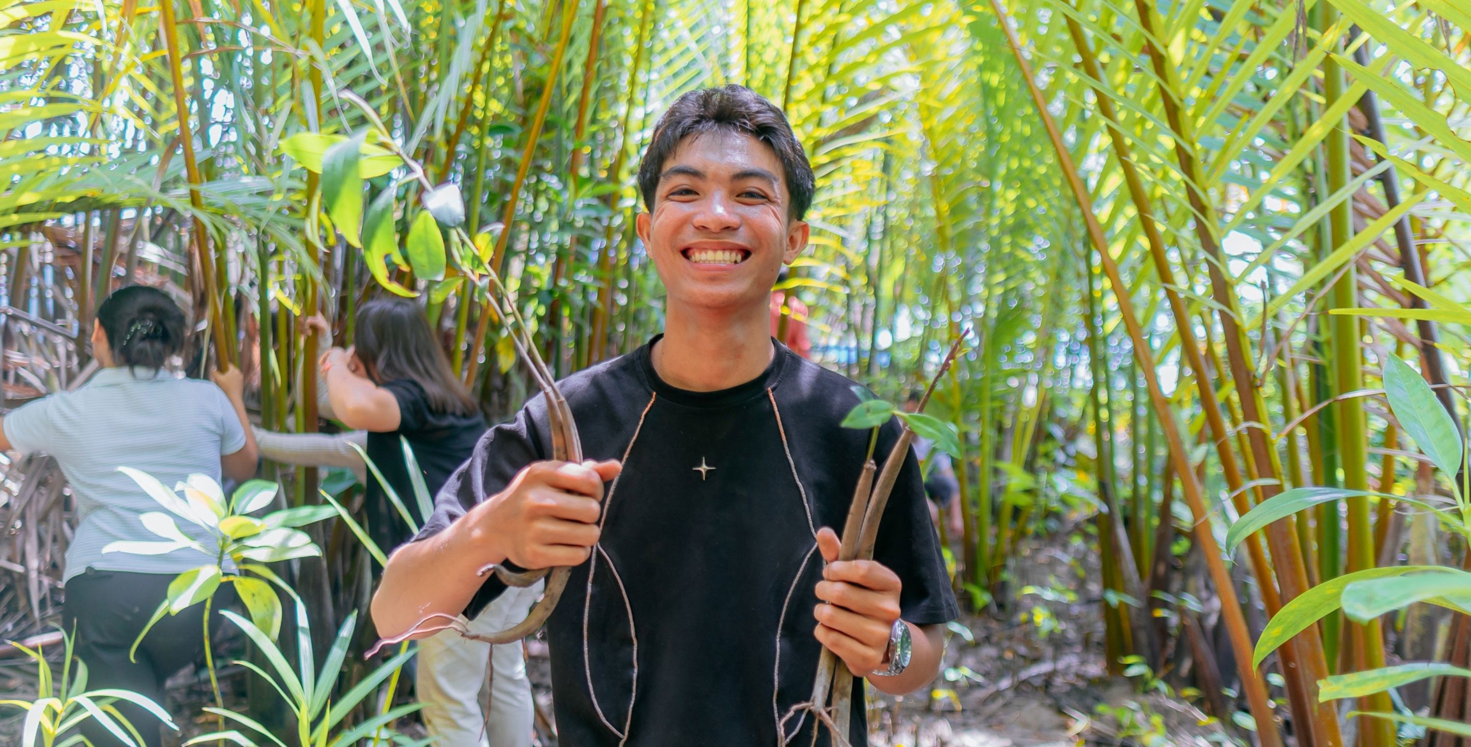 A young man in the Philippines stands in a mangrove forest, holding a couple branches and smiling widely.