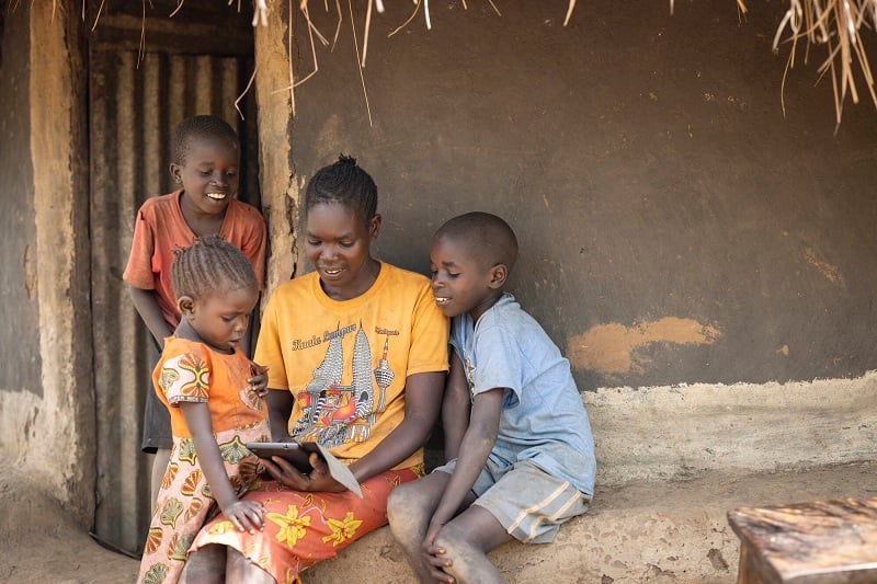 A mom in Uganda sits in front of a hut and plays on a tablet, surrounded by her smiling children.