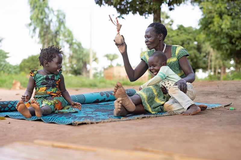 A grandmother plays with a homemade doll on a picnic blanket with her 2 grandchildren.