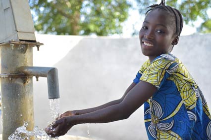 Girl stands outside at water well washing hands smiling in Sierra Leone.