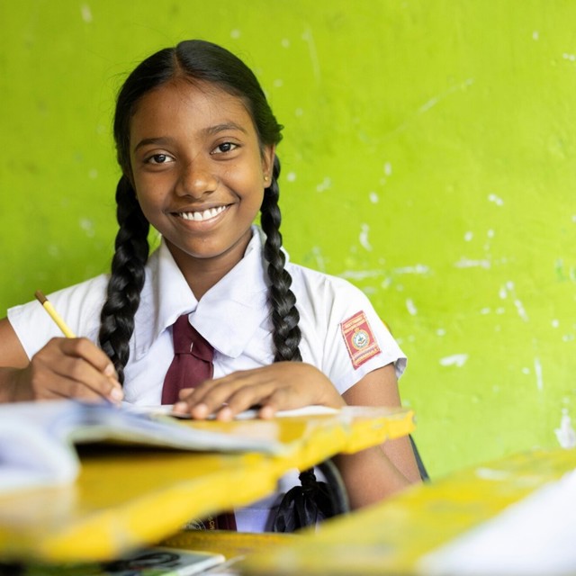 A young girl in a classroom smiles while holding a pencil against a vibrant green wall.