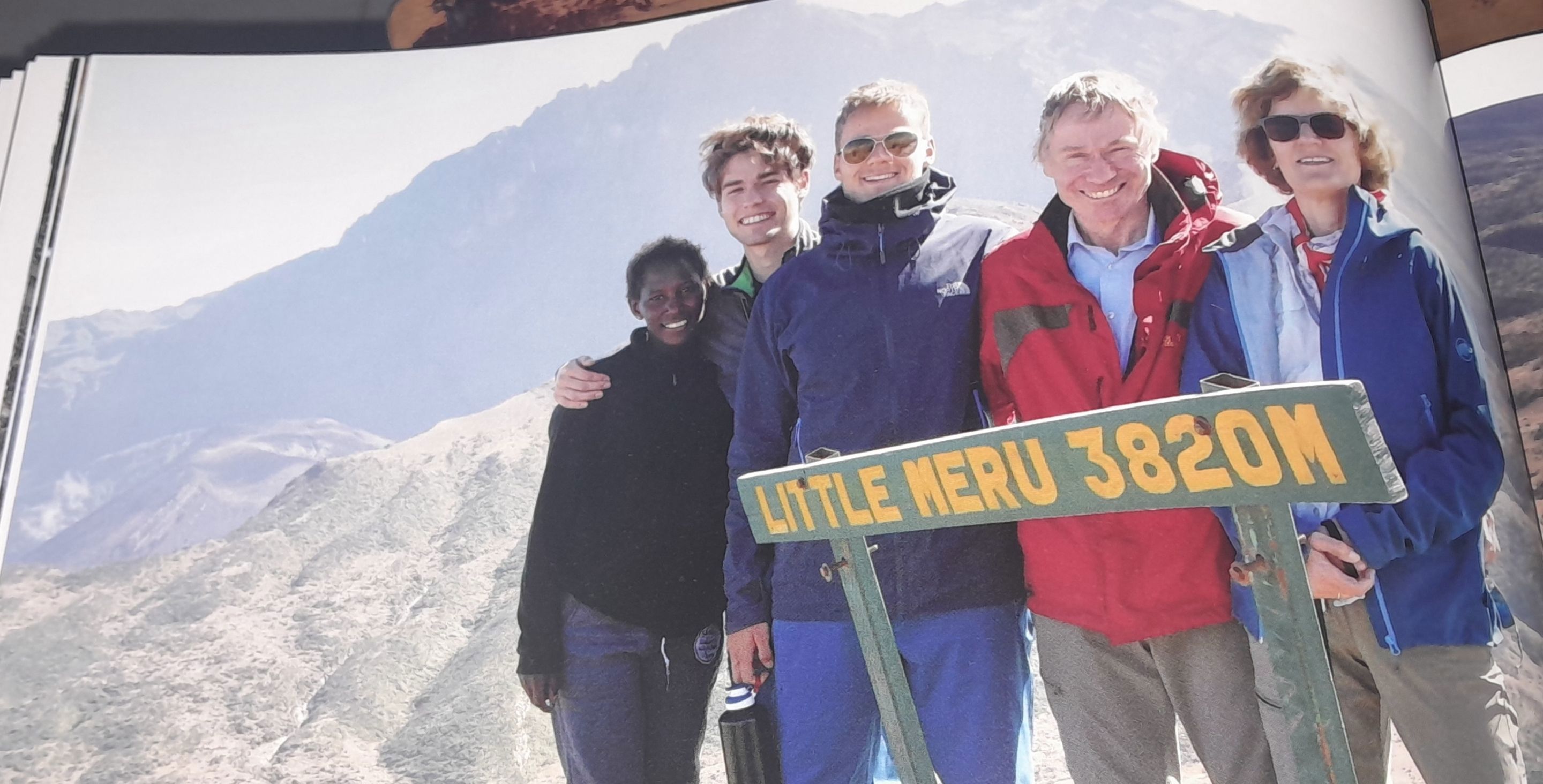 A family stands on top of a mountain smiling. The sign in front of them reads 