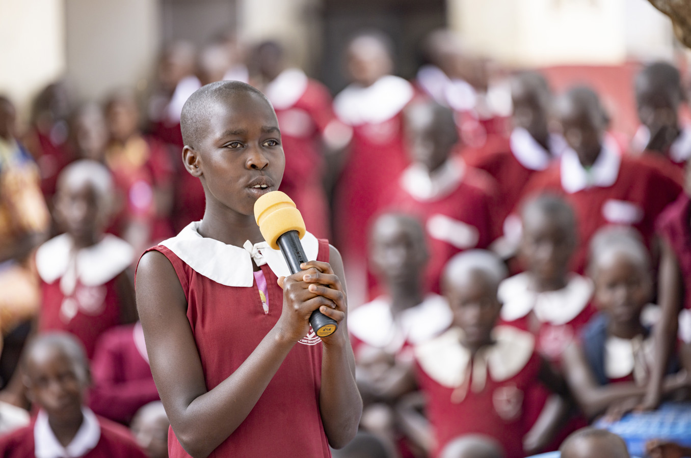A child in Uganda stands with a microphone in front of a group of other children.
