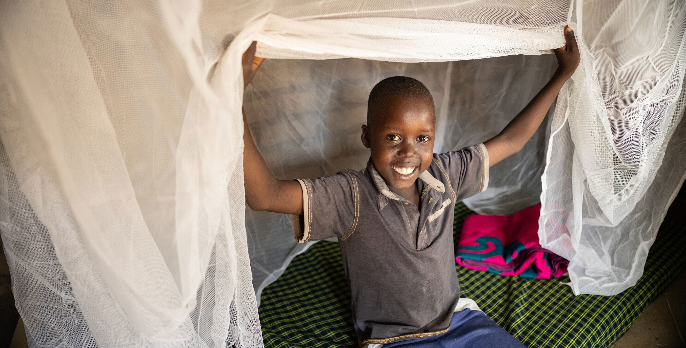 A little boy sits on a bed under a mosquito net, lifting the net up and smiling at the camera.
