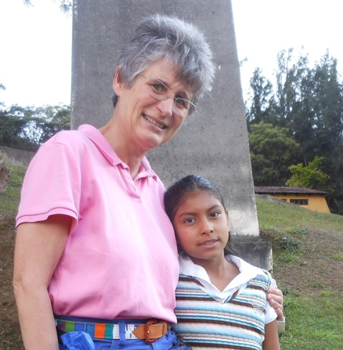 A woman and a girl pose together outdoors, smiling. The woman wears a pink shirt, and the girl has a striped sweater.