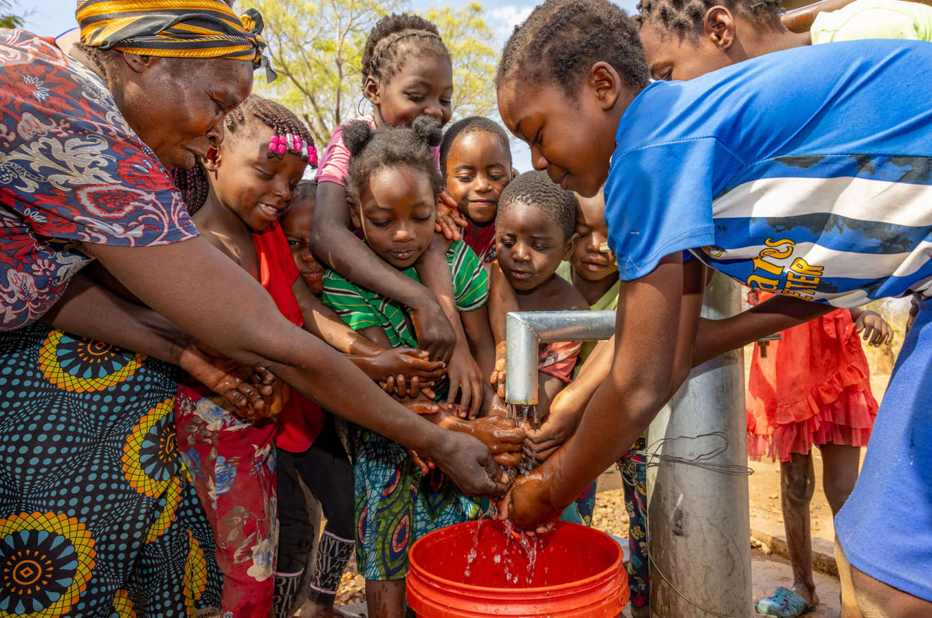 Children in Zambia gather around an outdoor water faucet, washing hands and smiling.