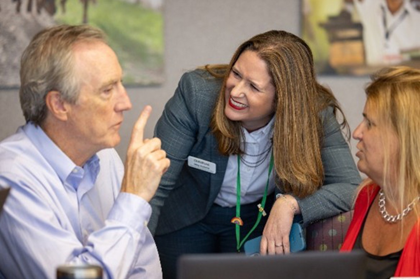 In an office meeting space, three individuals are gathered around a table. One person gestures with a raised finger while speaking, another leans in attentively, and a third sits beside them. Papers, laptops, and office decor are visible in the background, suggesting an active discussion or collaboration.