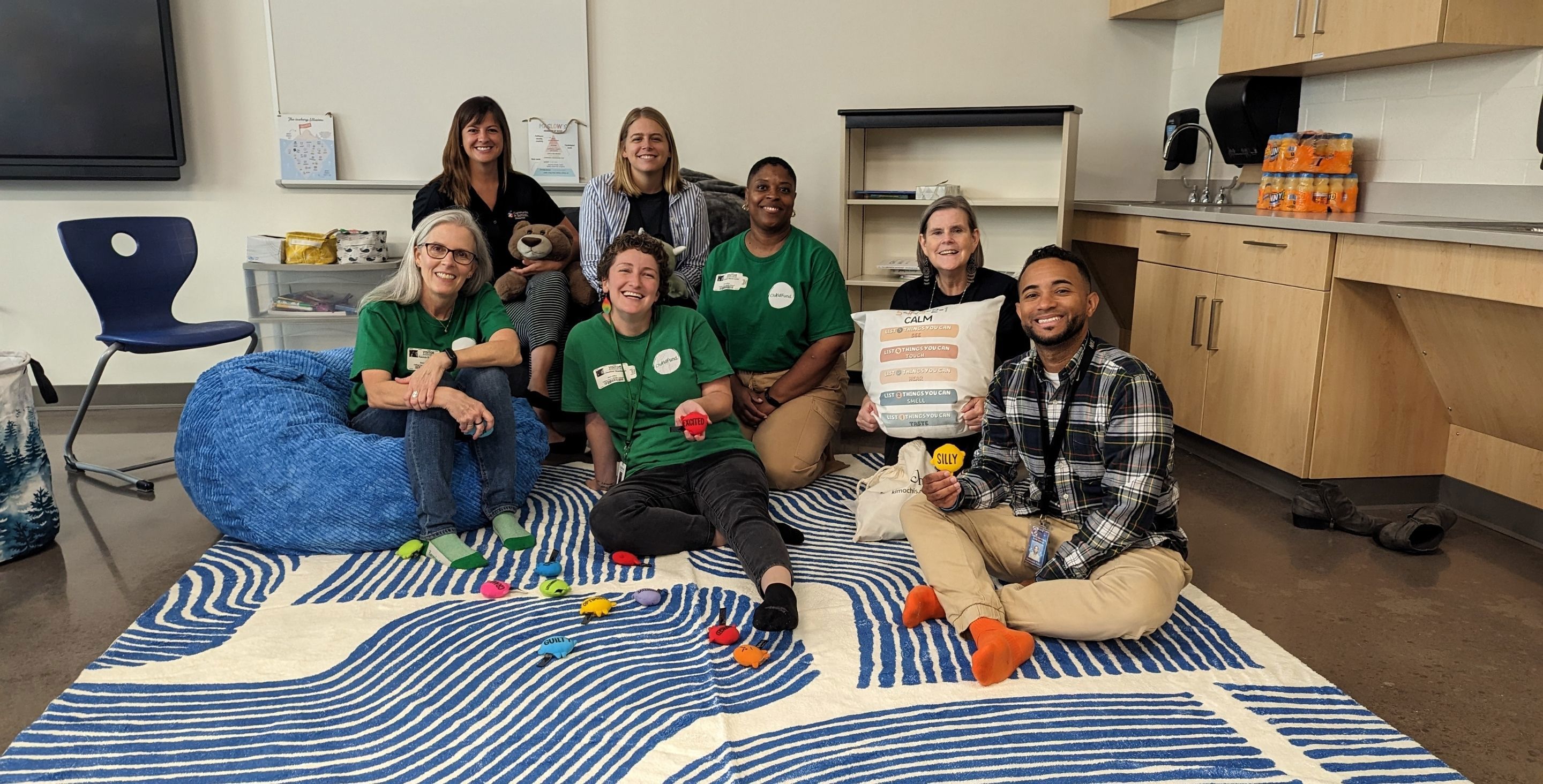 A group of adults, some wearing ChildFund shirts, sit on the floor in a classroom with warm lighting, books and bean bags.
