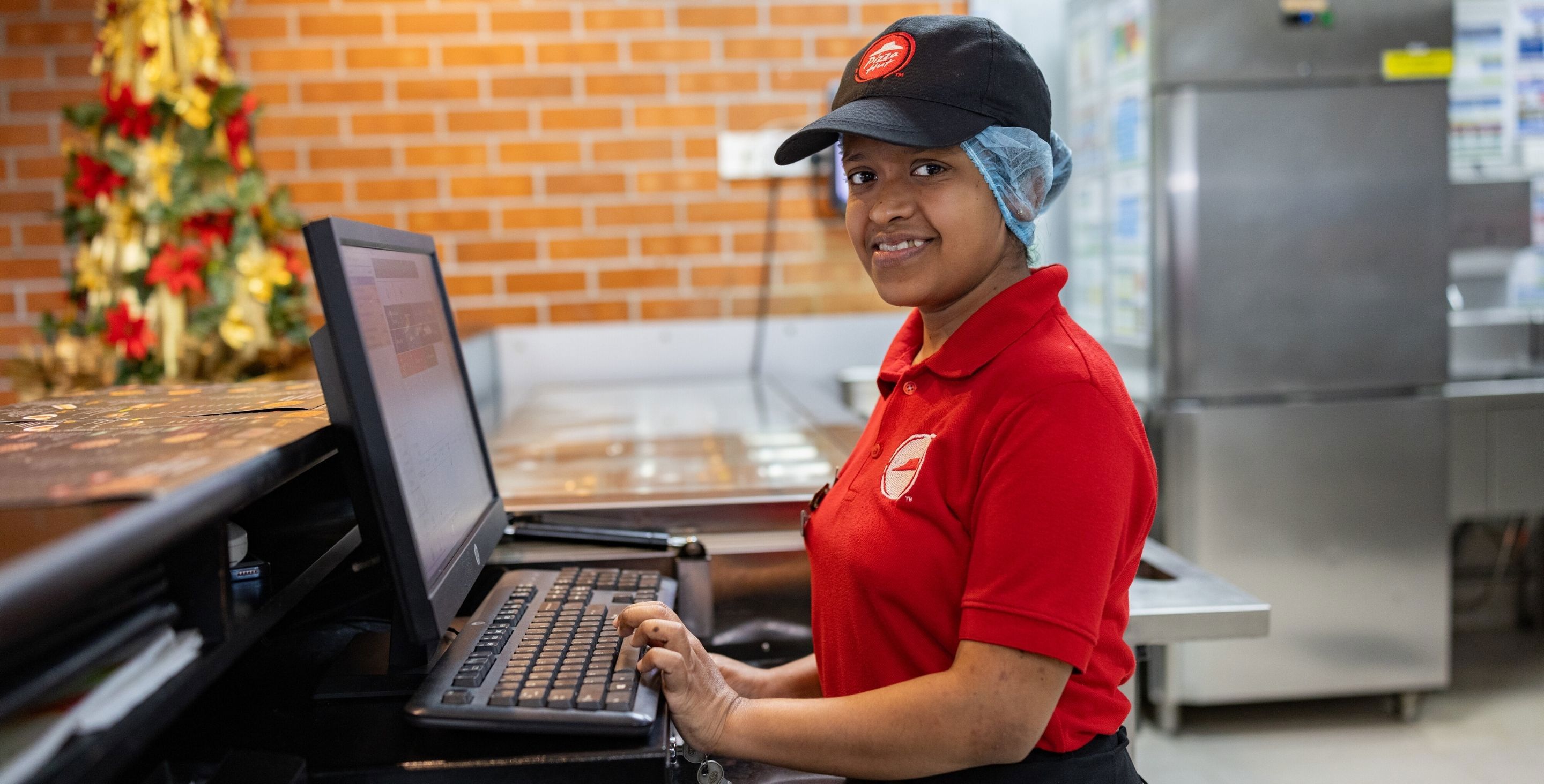 A young woman in Sri Lanka in a Pizza Hut uniform and a hair net stands at the cash register, smiling at the camera.