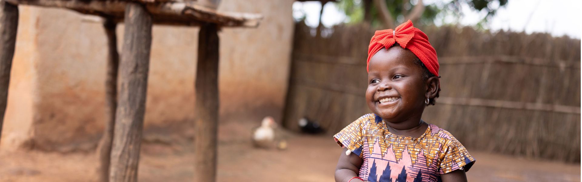 A little girl in Mozambique wearing a red head covering holds a homemade ball, smiling.