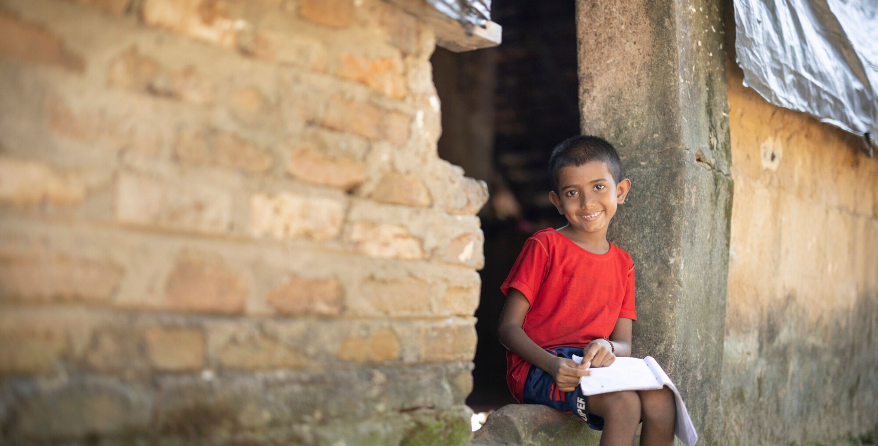 A sponsored child in Sri Lanka sits in the doorway of his earthen home, writing a letter to his sponsor.
