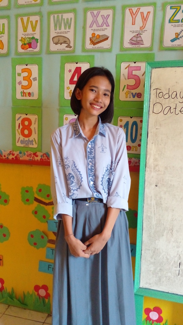 Teen girl in Indonesia stands in a colorful classroom, smiling at camera.