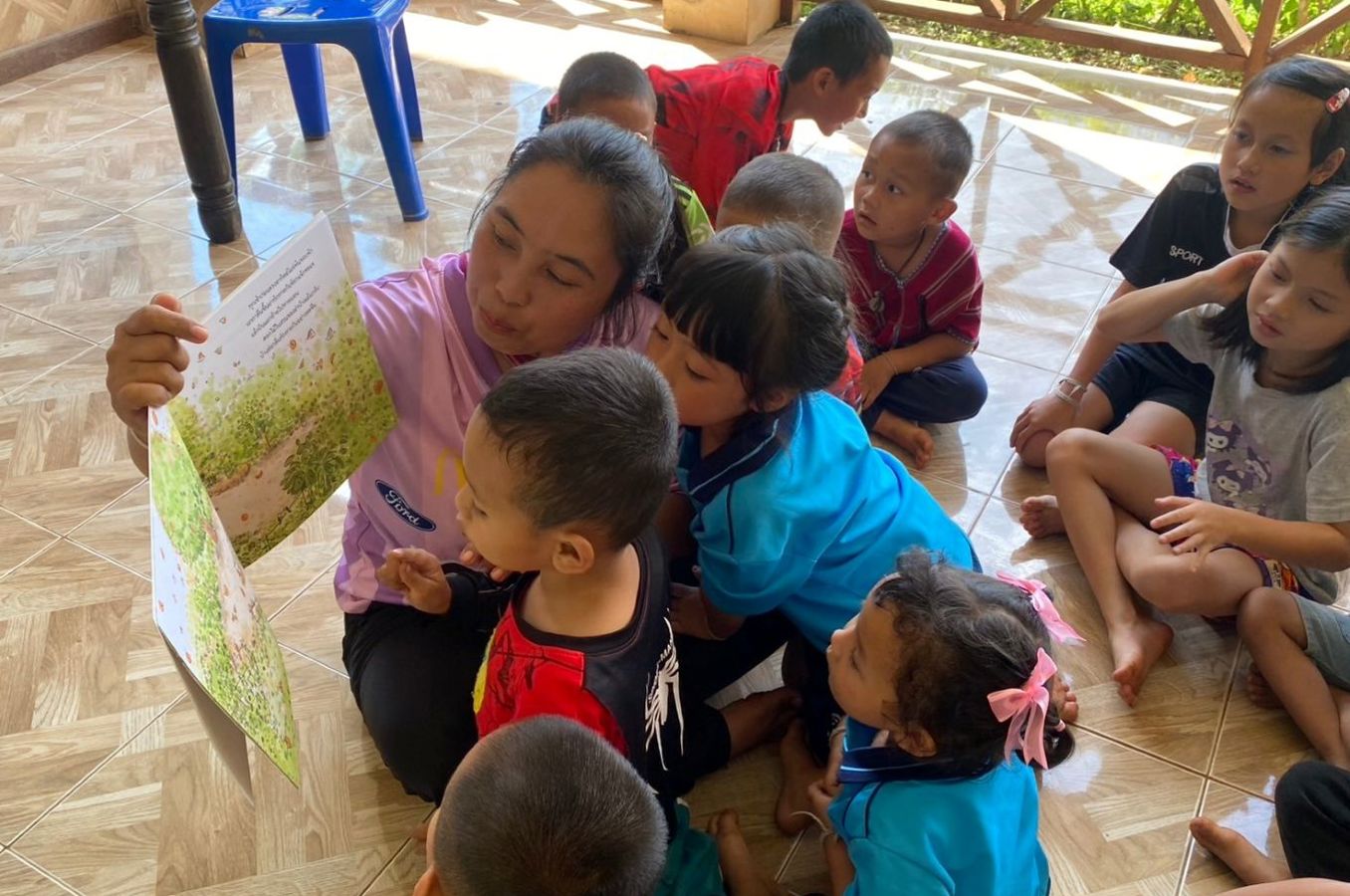 A woman sits on the floor in a classroom reading a book to a group of small children.