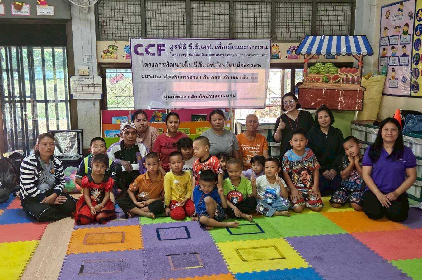 A group of young children and teachers gather on a colorful mat for a class picture. Above them is a sign that reads Community Children Foundation, with Thai writing beneath it.