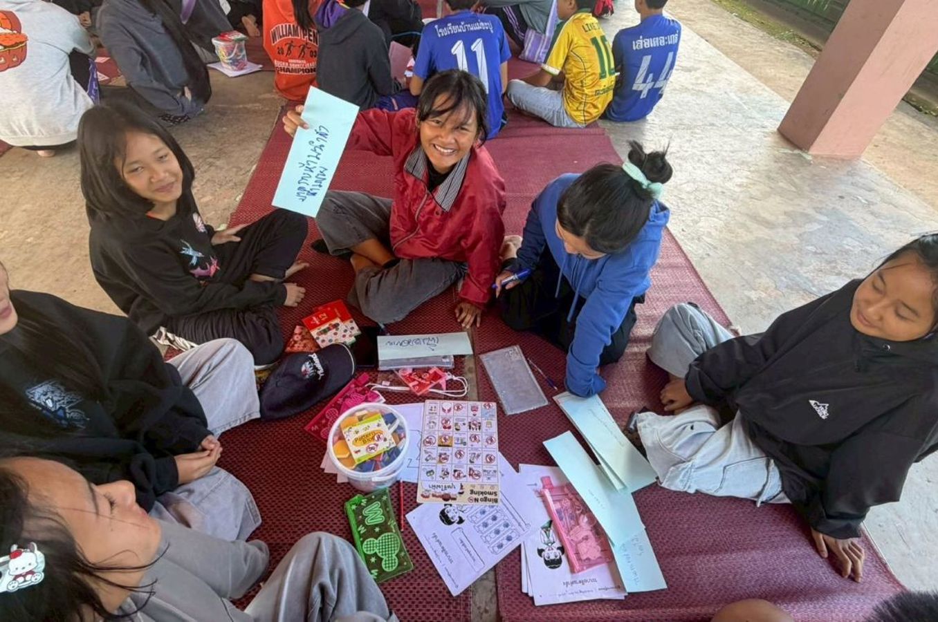 A group of teenagers sit on the floor in a circle, laughing together as they write on pieces of paper in the Thai language.