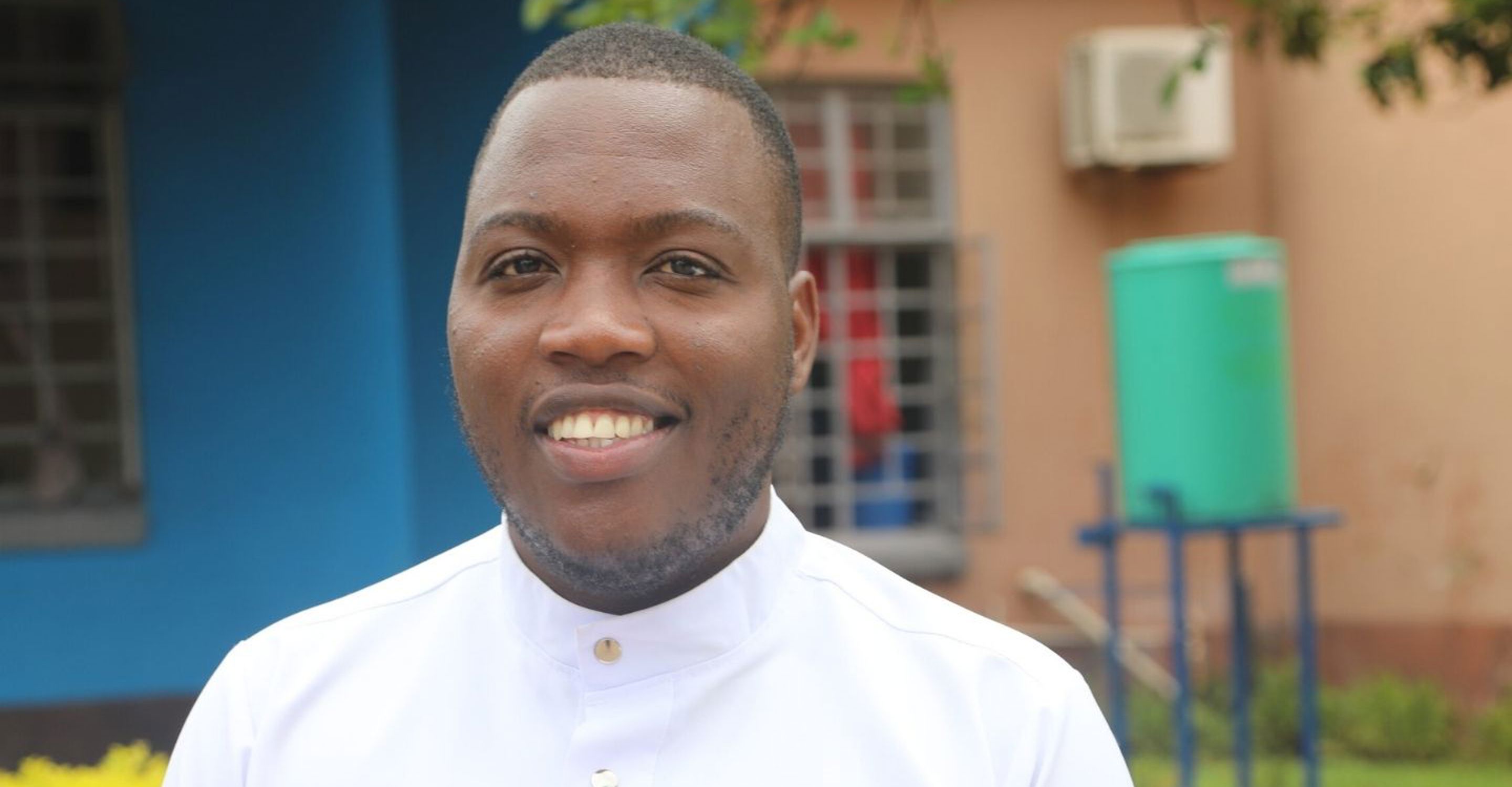 A young man wearing a nurse's uniform stands outside a hospital in Zambia, smiling at the camera.