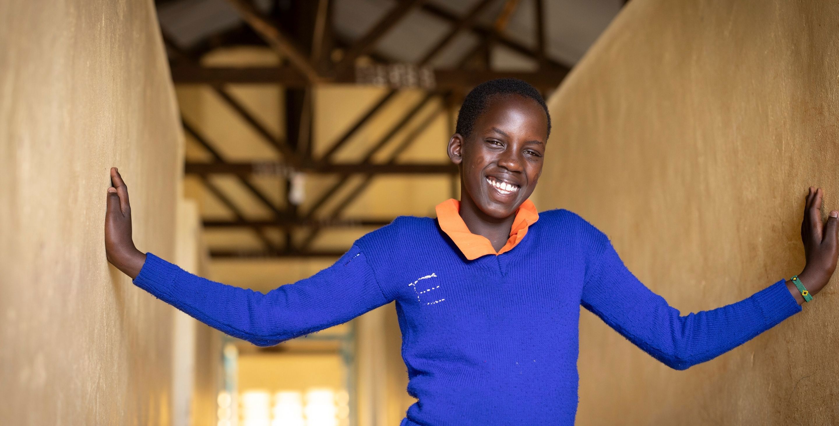 A girl in Kenya wearing a school uniform stands in the doorway of her doorway, arms outstretched, smiling.