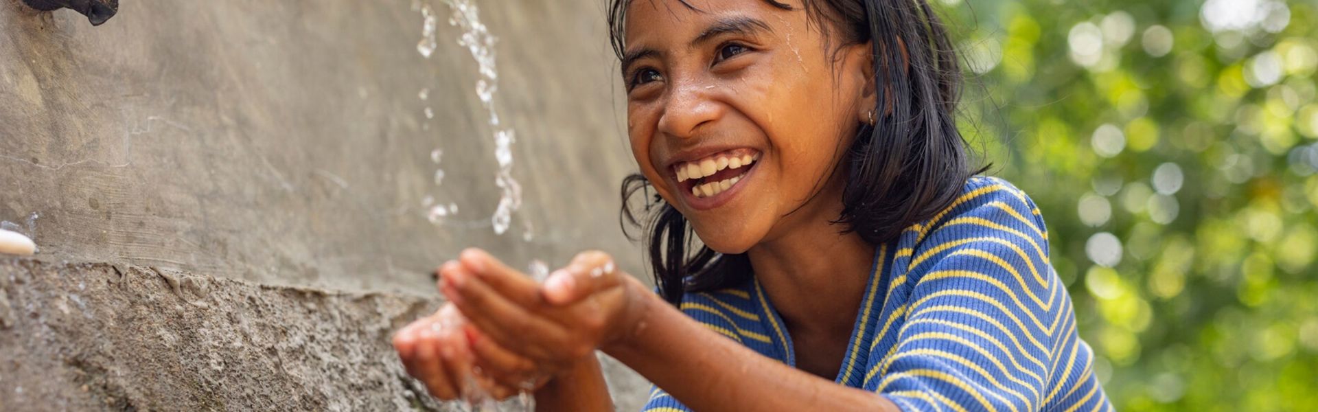 A girl in Indonesia wearing a striped shirt laughs while she washes her hands under an outdoor spigot.