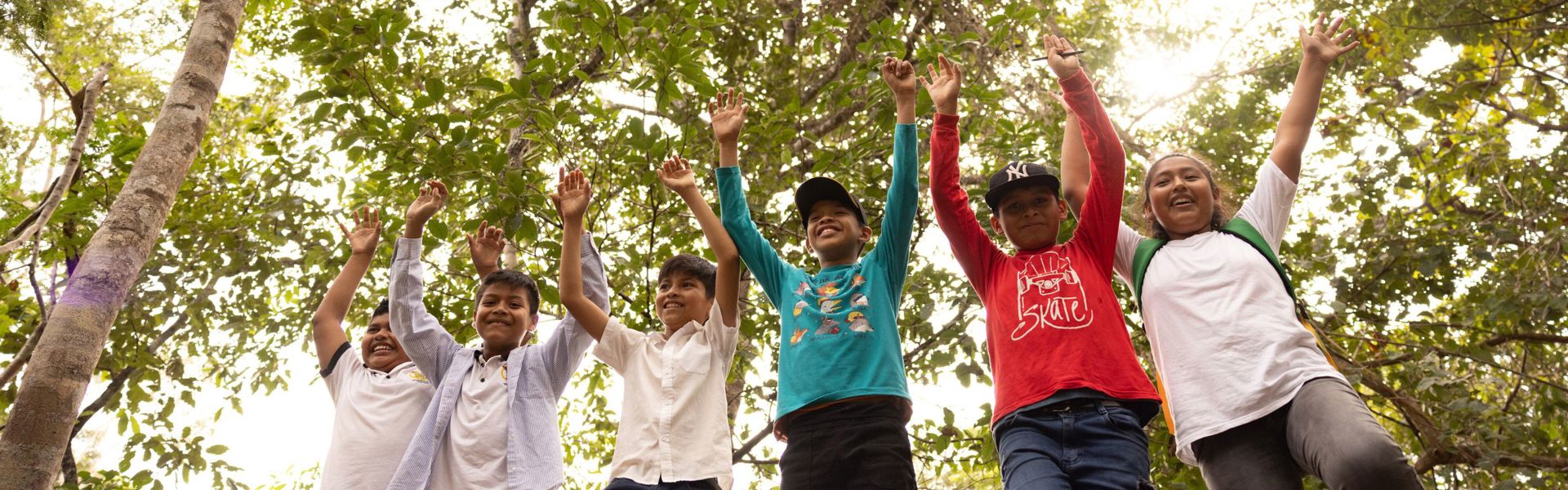 A group of children in Bolivia stand together in a forest, lifting their arms high and smiling.