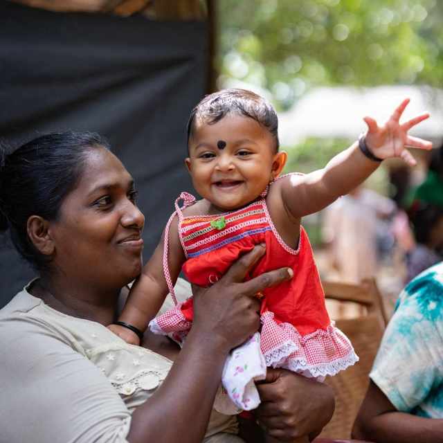 A mom holding up a very happy toddler girl, who is waving