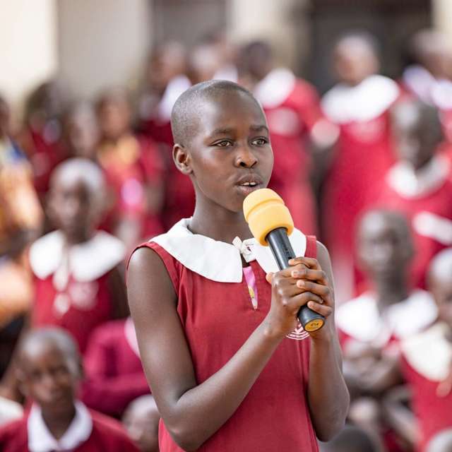 In Uganda, a 12-year-old girl in a school uniform holds a microphone to speak to her classmates about their rights.