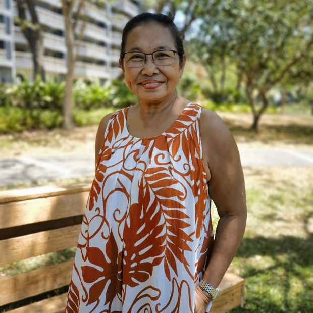 A Filipino woman wearing a Hawaiian print dress and glasses smiles at the camera, standing in a park