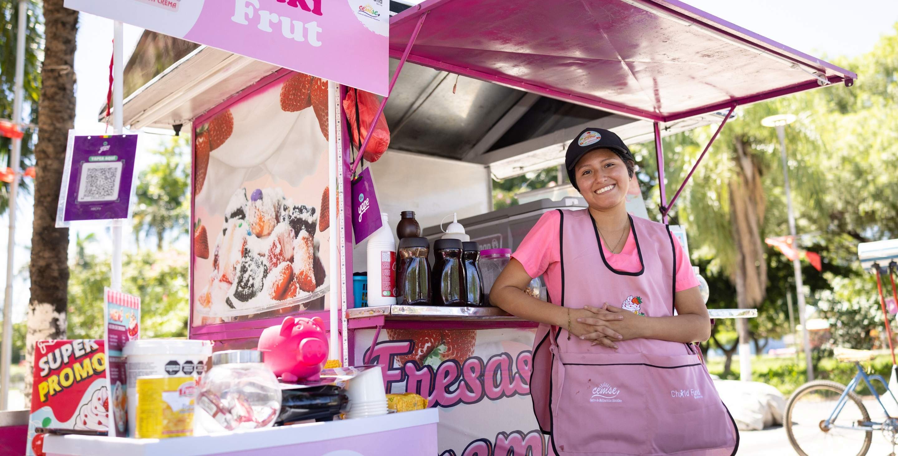 A young woman in Bolivia stands beside a dessert cart, smiling, wearing a pink apron.
