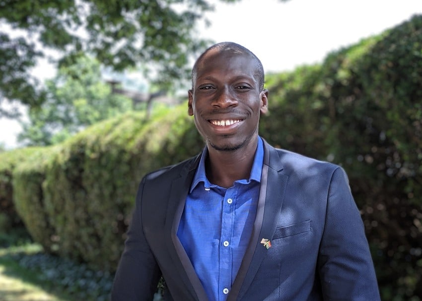 Portrait of young man from The Gambia standing outdoors smiling at the camera, wearing Gambia and United States pin.
