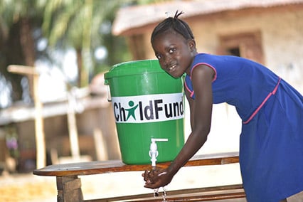 A 10-year-old girl in Sierra Leone washes her hands with water from a green bucket labeled ChildFund.
