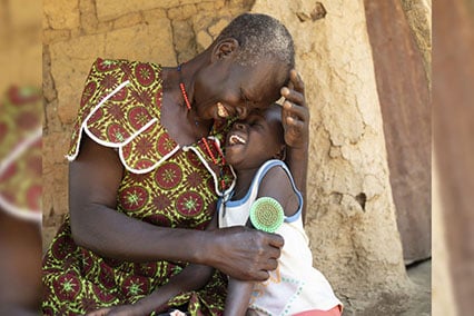A little girl and elderly woman hug and laugh outside a mud hut in Uganda.