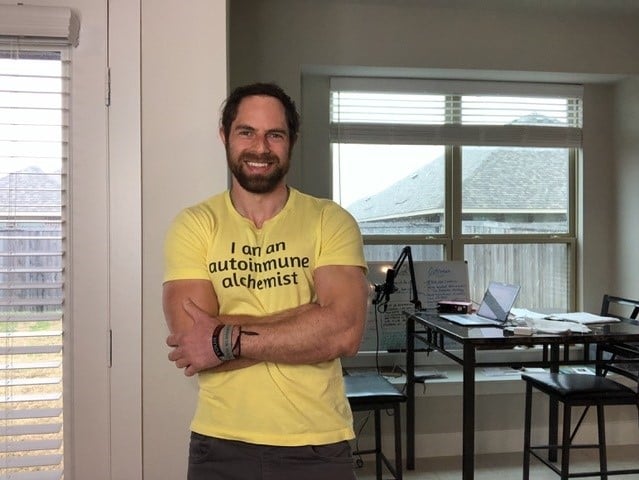 Man stands smiling at the camera wearing shirt that reads 'I am an autoimmune alchemist'.