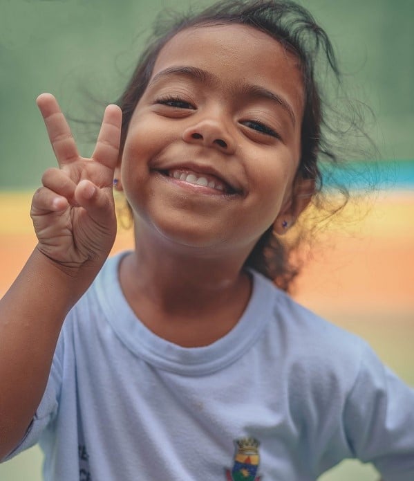 Girl in Brazil stands looking at camera and smiling, making a peace sign with her fingers.
