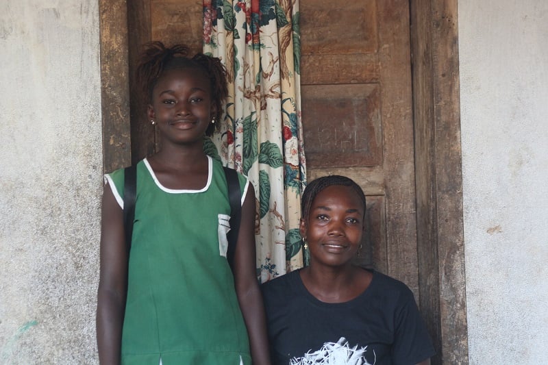 A mother and daughter sit in front of their house in Sierra Leone, looking at the camera.