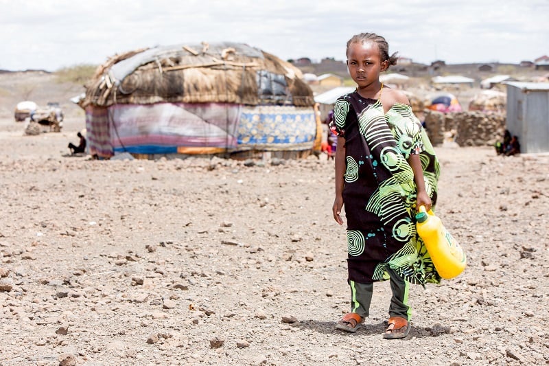 Young girl in desolate landscape in Kenya stares at camera, holding a jerrycan.