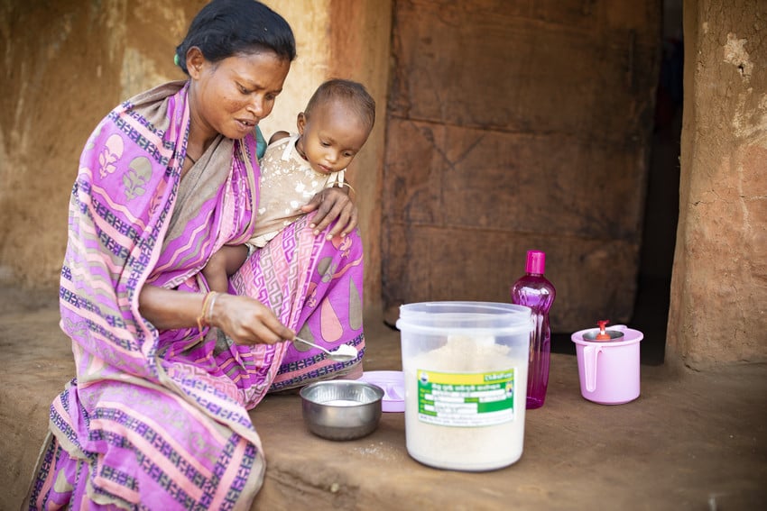 A mother sits on the porch of her mud hut in India with her small daughter, scooping a spoonful of dry porridge mix into a bowl.
