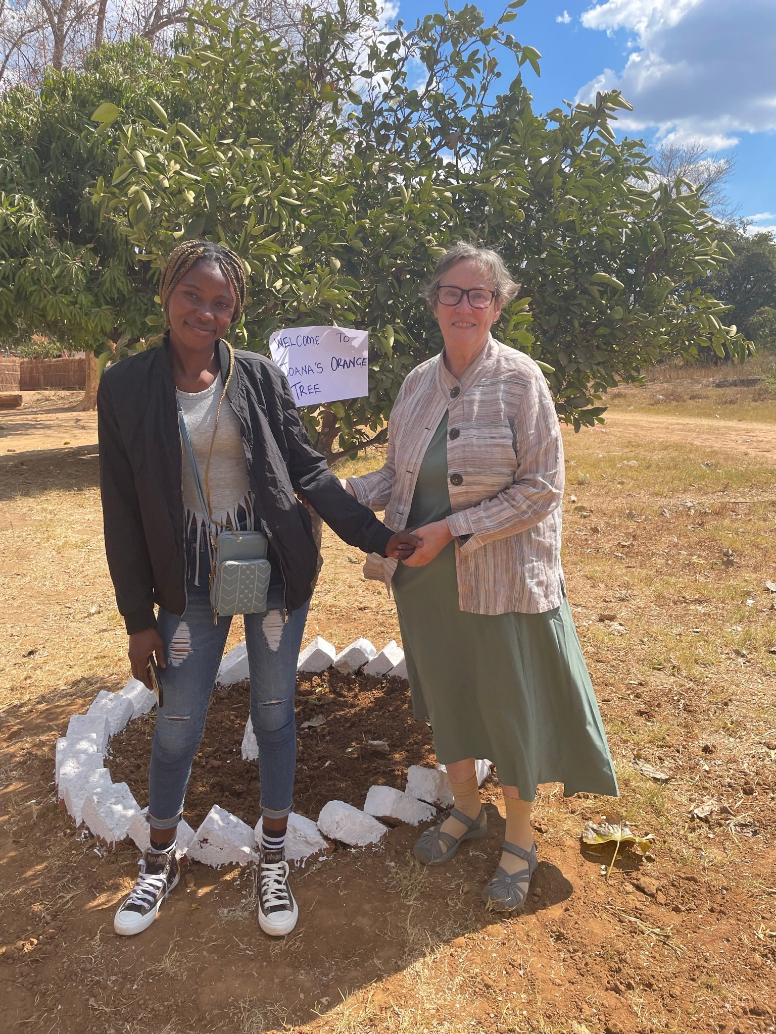Two women, one from Zambia and one from the U.S., stand in front of an orange tree.