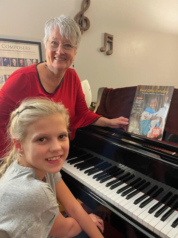 A piano teacher stands beside a young girl seated at the piano, with a copy of ChildFund's Real Gifts Catalog sitting on top of the piano.