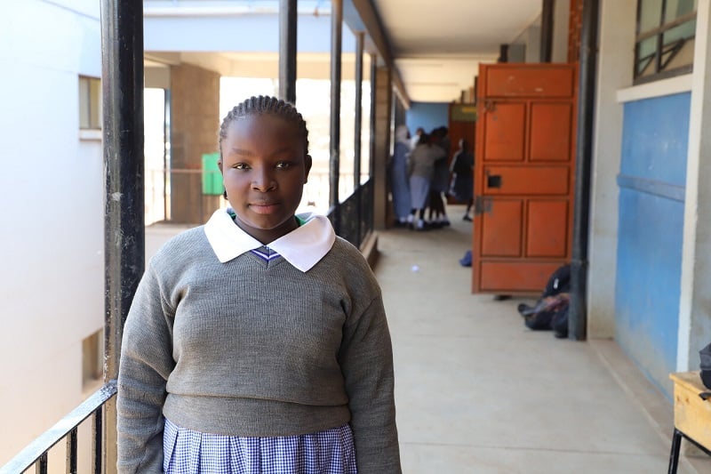A teenage girl in Kenya stands in a school hallway, looking seriously at the camera.