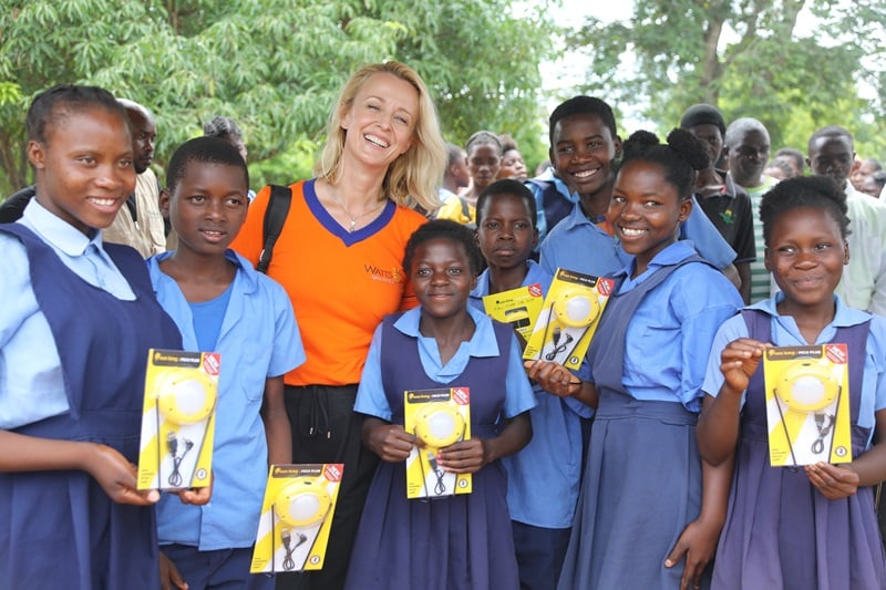 A woman stands with a group of children in Zambia holding up solar lamps