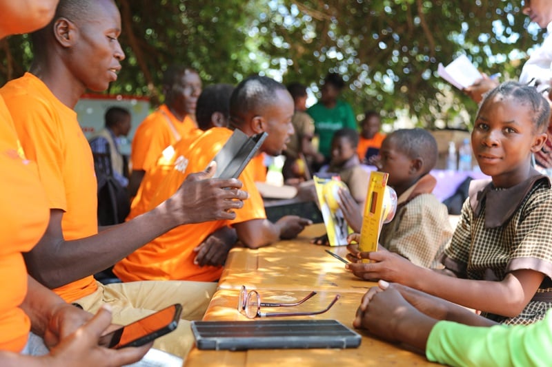 Children and parents line up at a table with people in orange shirts giving out solar lamps in Zambia