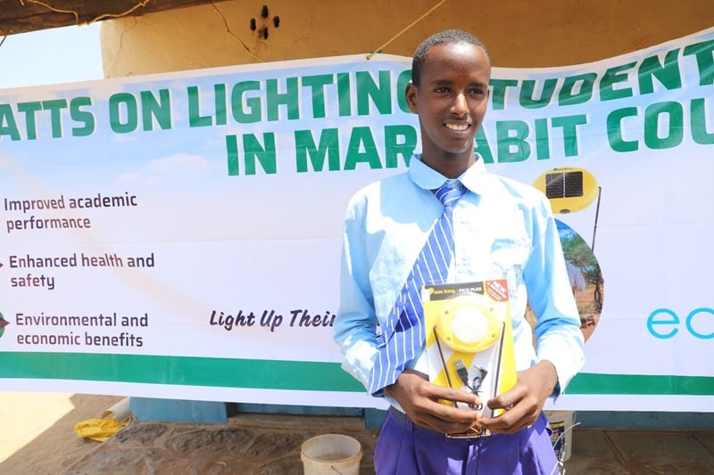 A young man smiles in front of a banner with the project name, holding up the solar lamp